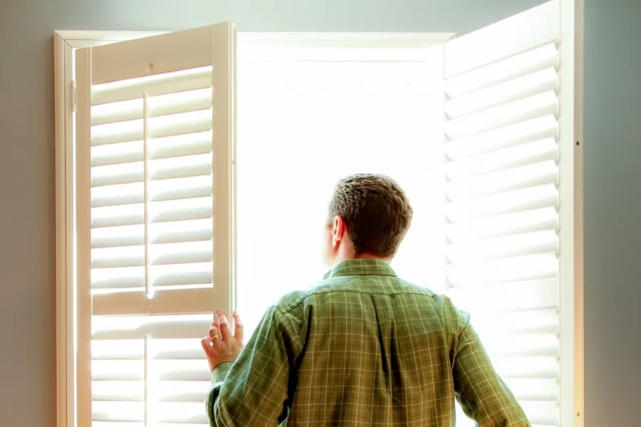 A man opening a pair of white plantation shutters onto bright daylight