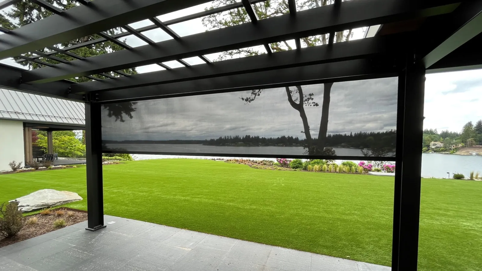 Exterior motorized shade lowered across a covered pergola looking out over a lawn to the water